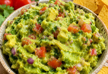Bol de guacamole maison frais avec morceaux d'avocat, coriandre et rondelles de piment, entouré de nachos croustillants sur une planche en bois
